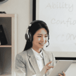 A smiling professional female Chinese teacher wearing headphones with a microphone, conducting a live 1-on-1 online Mandarin lesson with a tablet.